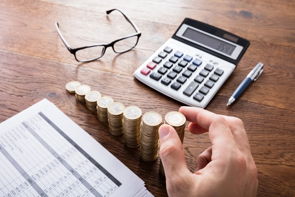 Mano apilando monedas junto a una calculadora y una planilla de cálculo, como al calcular las contribuciones de una propiedad