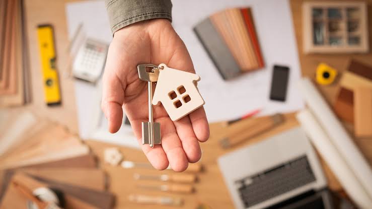 Mano sosteniendo las llaves de una propiedad con llavero en forma de casa, sobre un escritorio con planos y calculadora